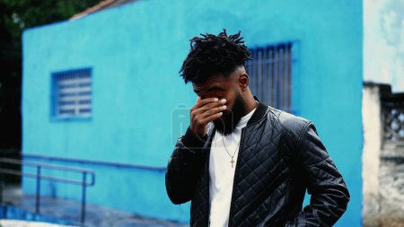 Téléchargez les photos : Un jeune homme noir fatigué debout dehors dans la rue de la ville touchant le visage et les yeux, vêtu d'une veste sous la pluie de bruine. 20s personne après une nuit difficile - en image libre de droit