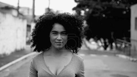 One confident young black woman walking towards camera outside in urban street environment in intense monochromatic, black and white. Curly hair African American 20s girl
