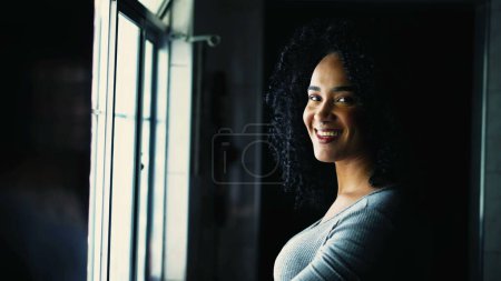 Foto de Feliz Retrato de una joven brasileña negra girando la cabeza hacia la cámara sonriendo de pie junto a la ventana en casa. Cabello rizado chica adulta sudamericana sonriendo - Imagen libre de derechos
