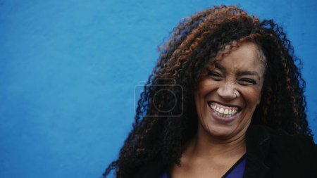Foto de Una mujer negra feliz de 40 años sonriendo y riéndose de la cámara en el fondo azul en la calle de la ciudad. Retrato de una persona afrodescendiente alegre en la vida urbana de América del Sur - Imagen libre de derechos