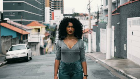 Photo for One confident young black woman with curly hair standing outside in urban setting looking at camera with stern solemn expression. South American lating 20s person in street portrait face - Royalty Free Image