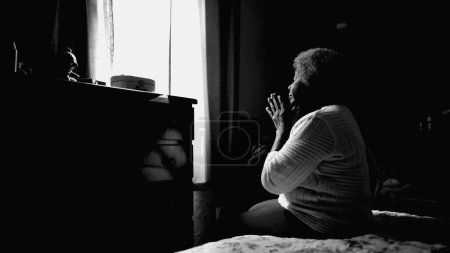 Foto de Una tranquila dama afroamericana de 80 años rezando en casa con los ojos cerrados. Una mujer negra esperanzada y agradecida en ORACIÓN en monocromática - Imagen libre de derechos