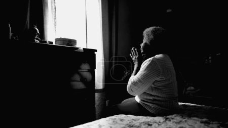 Téléchargez les photos : Une vieille afro-américaine tranquille dans les années 80 priant à la maison les yeux fermés. Une femme noire pleine d'espoir et reconnaissante en PRIERE en monochromatique - en image libre de droit