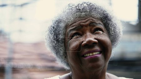 Esperanzada mujer afroamericana mayor mirando al cielo durante la lluvia con FE y ESPERANZA. Una dama negra de pelo gris girando la cabeza hacia arriba