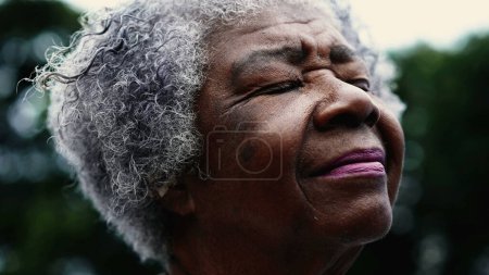 Téléchargez les photos : Reconnaissante Femme afro-américaine âgée fermant les yeux dans la contemplation spirituelle debout à l'extérieur, gros plan sur le visage d'une dame aux cheveux gris âgée noire dans les années 80 se sentant présence de DIEU - en image libre de droit