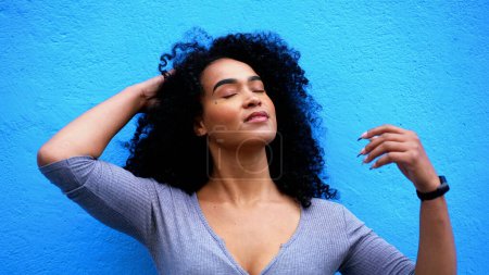 Foto de Una atractiva joven negra sonriendo a la cámara en un entorno urbano de pie en una pared azul. chica adulta de ascendencia africana de 20 años ajustando la sensación de cabello rizado empoderado y seguro - Imagen libre de derechos