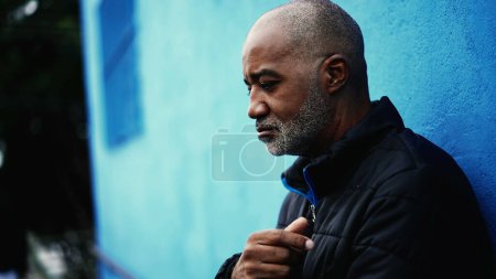 Téléchargez les photos : Un cher homme noir d'âge moyen se tenant dehors dans la rue de la ville regardant vers le haut dans une profonde expression contemplative. Regard réfléchi d'un Afro-Américain dans les années 50 - en image libre de droit