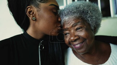 Caring granddaughter kisses elderly 80s grandmother in forehead in sign of deep love and support of inter-generational family moment. African American individuals