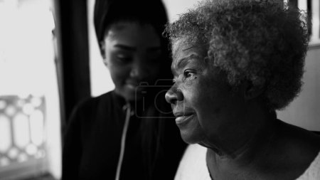 Pensive African American Senior woman staring at a distance with contemplative gaze, granddaughter in background showing support for grandmother in old 80s age, black and white