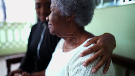 Téléchargez les photos : Petite-fille afro-américaine s'occupant de grand-mère âgée dans la vieillesse avec le bras autour de l'épaule montrant de l'aide et du soutien pour la femme âgée dans les années 80, soins familiaux intergénérationnels - en image libre de droit