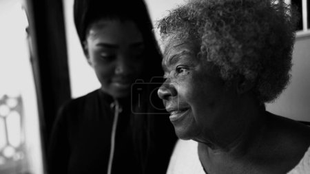 Umsichtige afroamerikanische Seniorin starrt mit nachdenklichem Blick in die Ferne, Enkelin im Hintergrund zeigt Unterstützung für Großmutter im Alter von 80, schwarz-weiß