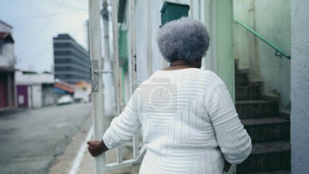Téléchargez les photos : Une dame noire âgée ouvre la porte d'entrée de la résidence, arrivant à la maison par la rue du trottoir urbain. 80 femmes afro-américaines retourne à la maison sud-américaine après une activité quotidienne de routine - en image libre de droit