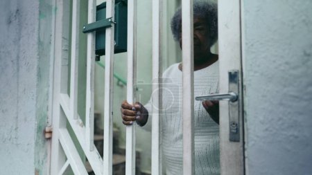 Téléchargez les photos : Une dame noire âgée ouvre la porte d'entrée de la résidence, arrivant à la maison par la rue du trottoir urbain. 80 femmes afro-américaines retourne à la maison sud-américaine après une activité quotidienne de routine - en image libre de droit