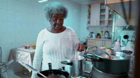 Téléchargez les photos : Seniors Femme afro-américaine âgée préparant le repas à la maison dans la scène de style de vie domestique décontractée en Amérique du Sud, un cheveux gris dame noire plus âgée cuisine - en image libre de droit