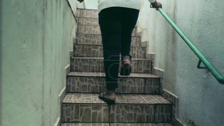 Photo for Back of humble active elderly African American senior woman going up the stairs arriving at residence in South America holding into metal bar on wall, bellow perspective - Royalty Free Image