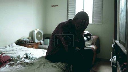 Foto de Un deprimido hombre negro de mediana edad sentado junto a la cama sufriendo una enfermedad mental en un dormitorio sombrío y malhumorado, de 50 años de edad, una persona afrodescendiente lucha contra la pobreza, cubriendo su rostro - Imagen libre de derechos