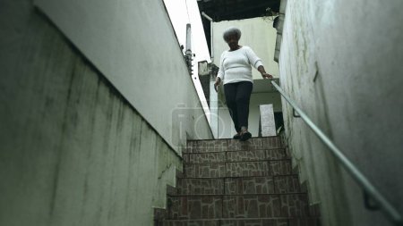 Foto de Una anciana negra sudamericana saliendo a la acera desde su residencia. 80s persona femenina de ascendencia africana bajando las escaleras y abre la puerta principal saliendo para la rutina diaria - Imagen libre de derechos