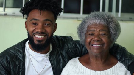 African American young grandson embracing his elderly gray hair grandmother portrait faces looking at camera in genuine loving moment, tender arm around shoulder, intergenerational bond