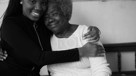 Photo for Tender loving moment of African American grandmother being held by teenage granddaughter in caring affectionate manner, portrait faces looking at camera smiling in black and white - Royalty Free Image