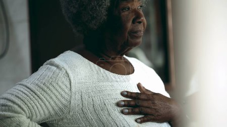 Téléchargez les photos : Une femme noire âgée réfléchissant à la maison, regardant par la fenêtre avec inquiétude et réflexion, main sur la poitrine - en image libre de droit