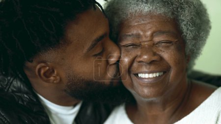 Téléchargez les photos : Petit-fils adulte embrassant une grand-mère afro-américaine âgée sur la joue dans un moment tendre et affectueux entre deux membres de la famille intergénérationnelle - en image libre de droit