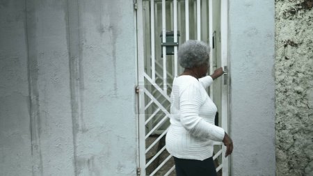 Photo for Elderly South American black woman arrives home from sidewalk street, opens residence front door returning by closing gate behind her, senior 80s person of African descent - Royalty Free Image