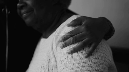 Téléchargez les photos : Gros plan de la main d'un membre de la famille autour de l'épaule d'une grand-mère âgée montrant de l'aide et du soutien pendant la vieillesse capturée en monochrome, noir et blanc - en image libre de droit