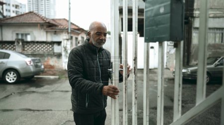 Téléchargez les photos : Un homme noir âgé descend les escaliers et ouvre la porte d'entrée de la résidence en Amérique du Sud, sortant dans le trottoir de la rue urbaine pendant la bruine. 50 personnes d'ascendance africaine quittant la maison - en image libre de droit