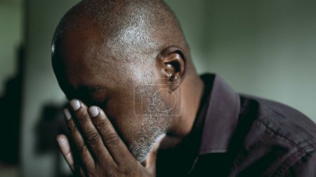 Photo for One middle-aged black 50s man in despair covers face with hands struggling with crisis. South American person in dimly lit room feeling desperation and loneliness during challening times - Royalty Free Image