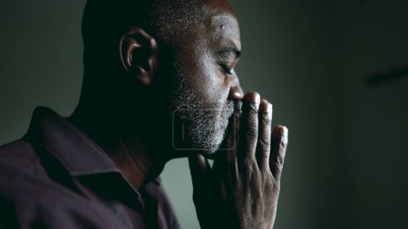Photo for One religious black senior man in contemplative PRAYER at home in dimly lit room with eyes closed having HOPE and FAITH during challenging times. Spiritual African American 50s person - Royalty Free Image