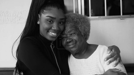 Photo for Tender loving moment of African American grandmother being held by teenage granddaughter in caring affectionate manner, portrait faces looking at camera smiling in black and white - Royalty Free Image