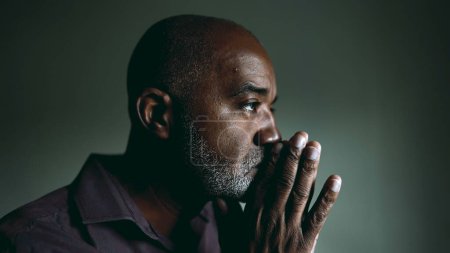 Téléchargez les photos : Un vieil homme religieux noir en prière contemplative à la maison dans une pièce faiblement éclairée avec les yeux fermés ayant l'ESPOIR et la Foi pendant les périodes difficiles. Spirituel afro-américain des années 50 personne - en image libre de droit