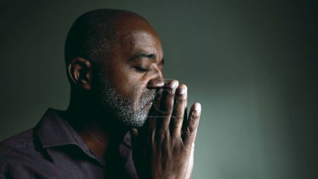 One religious black senior man in contemplative PRAYER at home in dimly lit room with eyes closed having HOPE and FAITH during challenging times. Spiritual African American 50s person