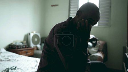 Téléchargez les photos : Un homme noir d "âge moyen déprimé assis au chevet d'un malade mental dans une chambre sombre et maussade, une personne d'ascendance africaine qui lutte contre la pauvreté, couvrant son visage - en image libre de droit