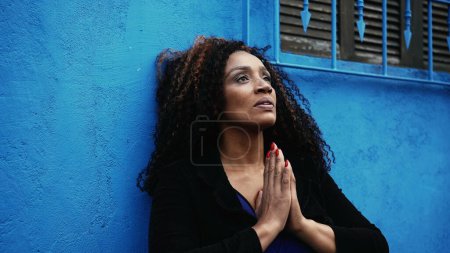 One worried hispanic black woman seeking solace during hard times Praying to GOD in urban setting gazing upwards with HOPE and FAITH