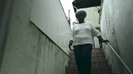 Photo for One elderly black lady from South American stepping out into sidewalk from residence. 80s female person of African descent going down the stairs and opens front door going out for daily routine - Royalty Free Image