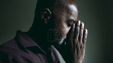 Téléchargez les photos : Un vieil homme religieux noir en prière contemplative à la maison dans une pièce faiblement éclairée avec les yeux fermés ayant l'ESPOIR et la Foi pendant les périodes difficiles. Spirituel afro-américain des années 50 personne - en image libre de droit
