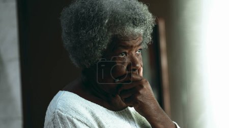 Photo for Pensive African American elderly woman lost in thought, standing by window gazing out in contemplative quiet preoccupation, old age depiction - Royalty Free Image