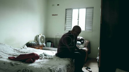 One depressed middle-aged black man seated in bedside suffering with mental illness in moody gloomy bedroom, 50s person of African descent struggles with poverty, covering face