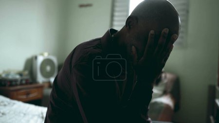 Photo for One depressed middle-aged black man seated in bedside suffering with mental illness in moody gloomy bedroom, 50s person of African descent struggles with poverty, covering face - Royalty Free Image