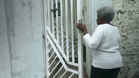 Téléchargez les photos : Femme noire sud-américaine âgée arrive à la maison de la rue du trottoir, ouvre la porte d'entrée de la résidence en revenant en fermant la porte derrière elle, personne âgée des années 80 d'origine africaine - en image libre de droit