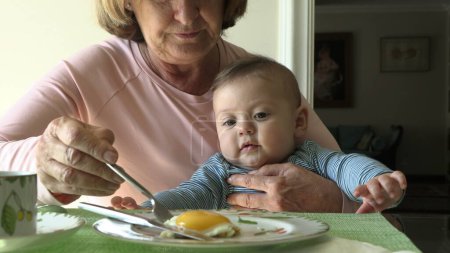 Téléchargez les photos : Grand-mère tenant petit-fils dans les bras pendant le petit déjeuner - en image libre de droit