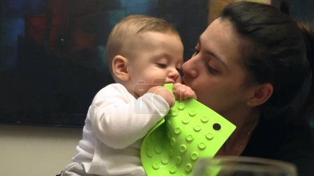 Téléchargez les photos : Bébé mettre un objet en plastique dans la bouche à la découverte du monde maman embrasser fils dans la joue occasionnel - en image libre de droit