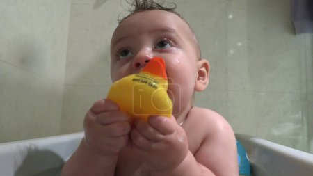 Téléchargez les photos : Mignon adorable bébé à l'intérieur baignoire mettre jaune canard dans la bouche - en image libre de droit