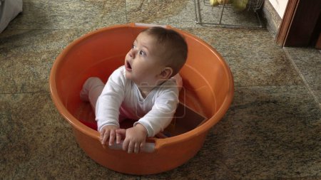 Téléchargez les photos : Adorable petit bébé tout-petit à l'intérieur grand seau à linge en plastique à la cuisine - en image libre de droit