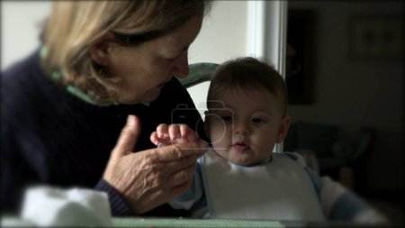 Téléchargez les photos : Casual franc authentique petit-fils bébé avec grand-mère à la table du déjeuner - en image libre de droit