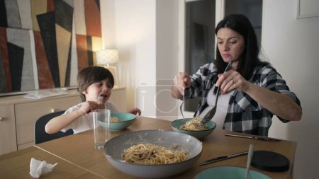 Foto de Cena juntos - Madre sirviendo pasta de espaguetis en el plato mientras su hijo come comida, alimentando el vínculo de la hora de la comida en casa - Imagen libre de derechos