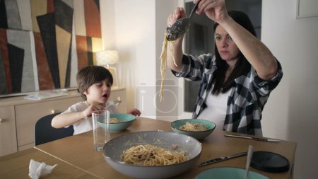 Dinner Together - Mother Serving Spaghetti Pasta on plate while son eats food, Nurturing Mealtime Bond at home