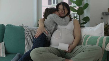Photo for Caring 5 year osd son hugging pregnant mother during third trimester of pregnancy, child and mom posing for camera seated on couch at home, mom holding ultrasound picture - Royalty Free Image