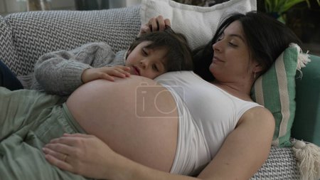 Photo for Maternal Bonding Moments - Young Son Expressing Love by Leaning on Expecting Mother's Belly, Relaxed Pregnant Mom Lounging on Sofa in Home Setting, Anticipating Baby's Arrival - Royalty Free Image